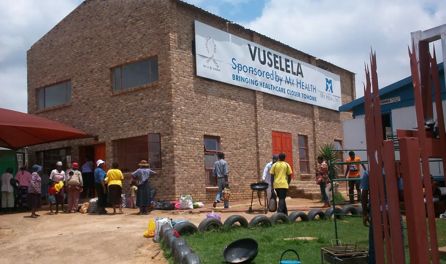 People gathered outside a brick building with a Vuselela sign.